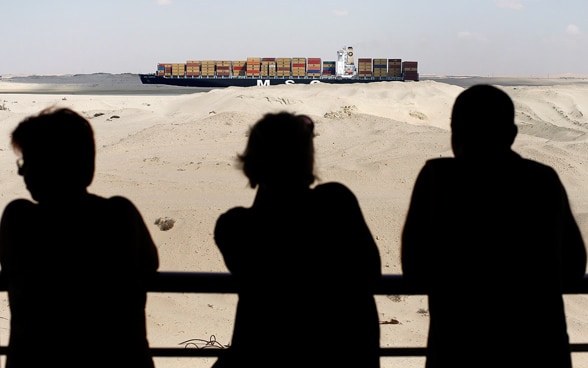Three people watch a boat carrying containers.