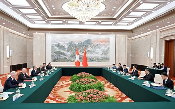 In a hall, the flags of Switzerland and China can be seen in the background. To the right and left of the room, delegations from the two countries are seated at a table.