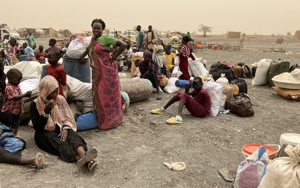 Women, children and young people who have fled Sudan wait with their belongings in a desert landscape.