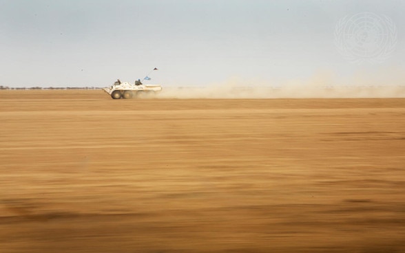 A white armoured vehicle of a UN peacekeeping mission drives at high speed through a desert landscape.