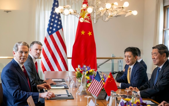 The US and Chinese delegations are seated at a wooden table. The flags of both countries can be seen in the background. 