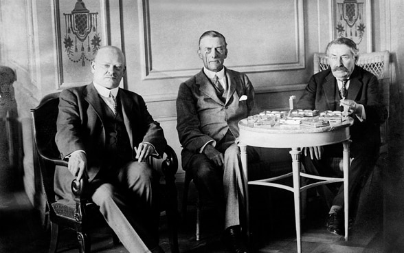 Schwarzweissaufnahme von Gustav Stresemann, Austen Chamberlain und Aristide Briand, die nebeneinander an einem Tisch sitzen.
