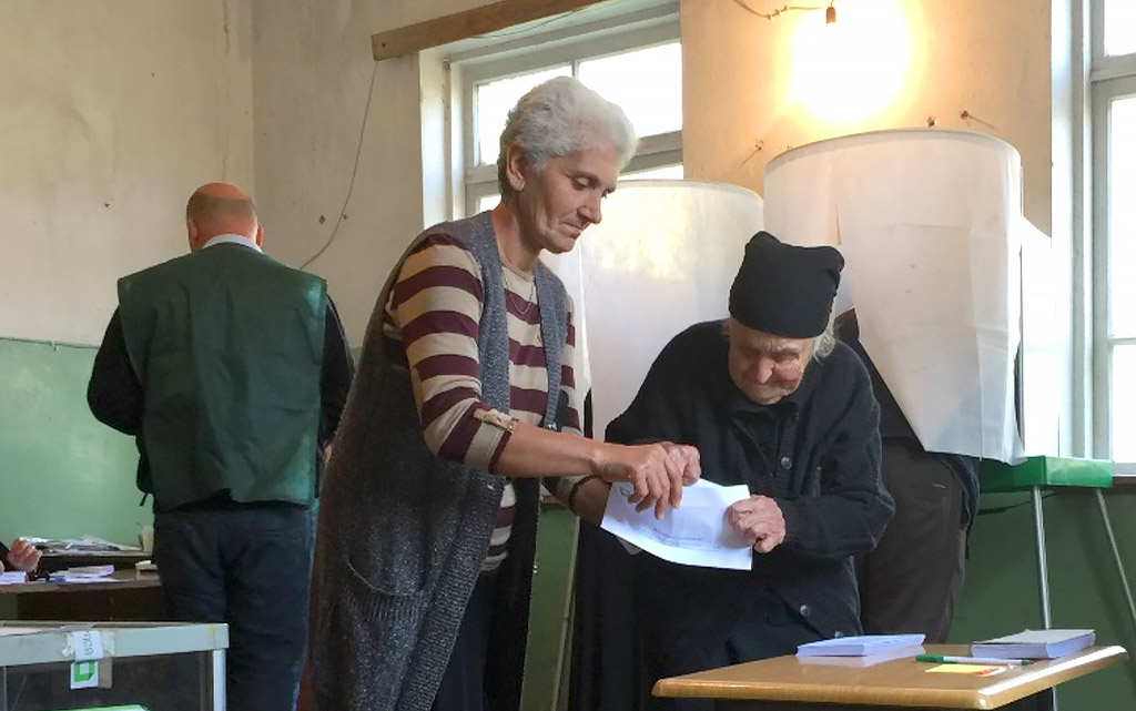 Deux femmes devant une urne lors des élections locales en Géorgie. L'une aide l’autre à plier un bulletin de vote dans un bureau de vote.