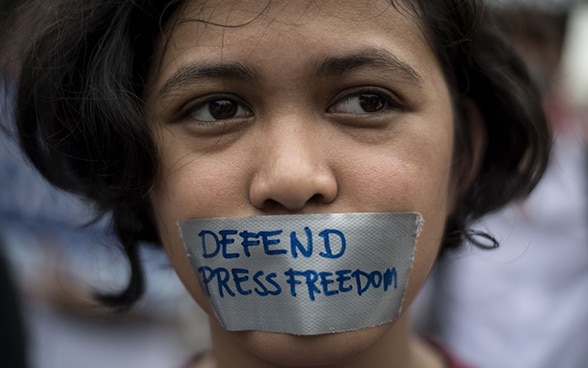 Portrait photo of a young girl. Her mouth is covered with a piece of duct tape, with the words 'defend press freedom' written on it.