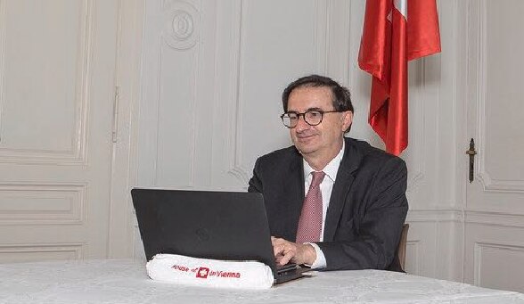 Botschafter Wolfgang Amadeus Brülhart sitzt im Home-Office und arbeitet an seinem Laptop. Hinter ihm ist eine Schweizer Fahne zu sehen.