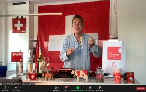 Major Thomas Schmidt preparing raclette in a room decorated with Swiss motifs. The scene was live-streamed using Zoom.