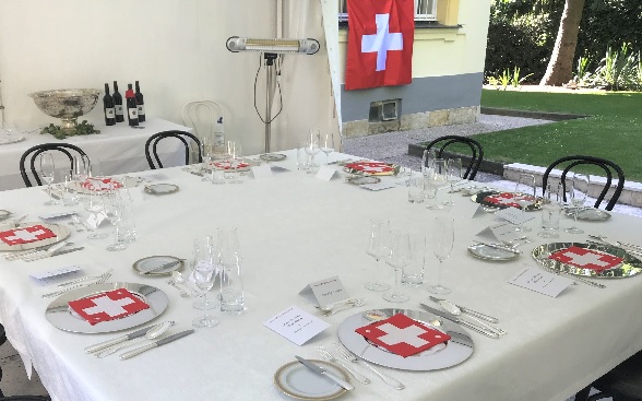 A table laid for eight persons under a canopy in the residence garden. On the plates are napkins decorated with images of the Swiss cross while a Swiss flag hangs in the background.