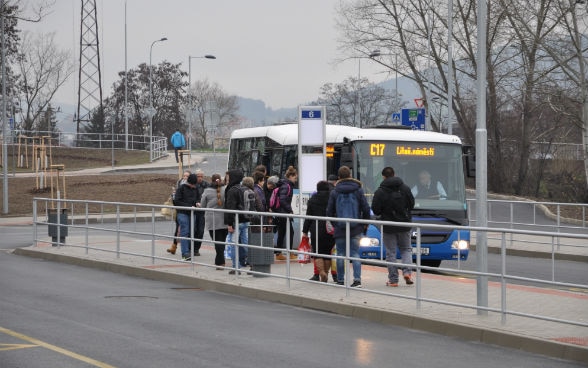 Un piccolo gruppo di persone sale su un autobus locale alla nuova stazione degli autobus. Alberi appena piantati e zone verdi testimoniano dell’impegno per rivitalizzare il territorio. 