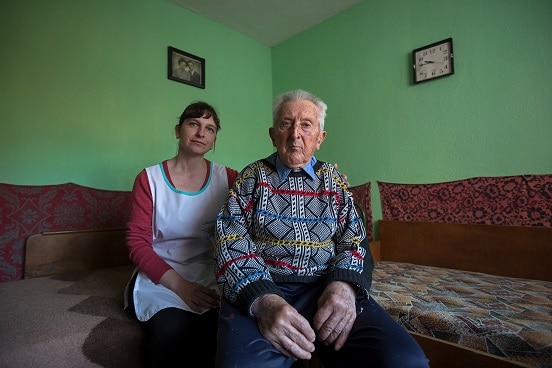 Un homme âgé, assis sur un canapé dans sa maison, en compagnie d’une aide-soignante.