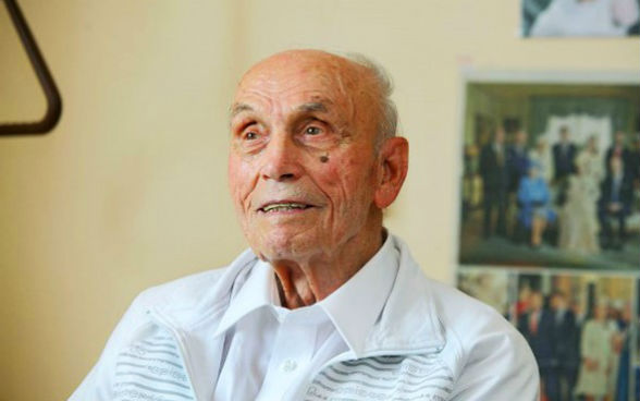 An old man wearing a white shirt and jersey sitting in a room.