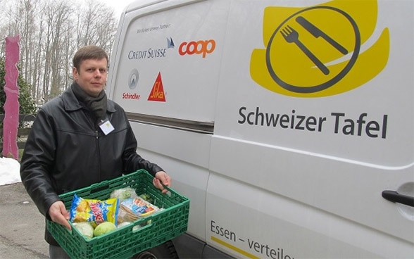 A man holds a crate of food in front of a van displaying the “Schweizer Tafel” and Coop logos.